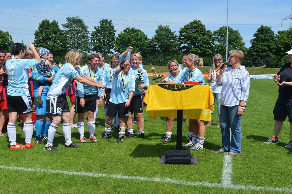 F&uuml;nfzehnfu&szlig;ballspielerinnen feiern um einen Tisch mit einer Fahne, w&auml;hrend ein Mitglied des Organisationsteams l&auml;chelt.