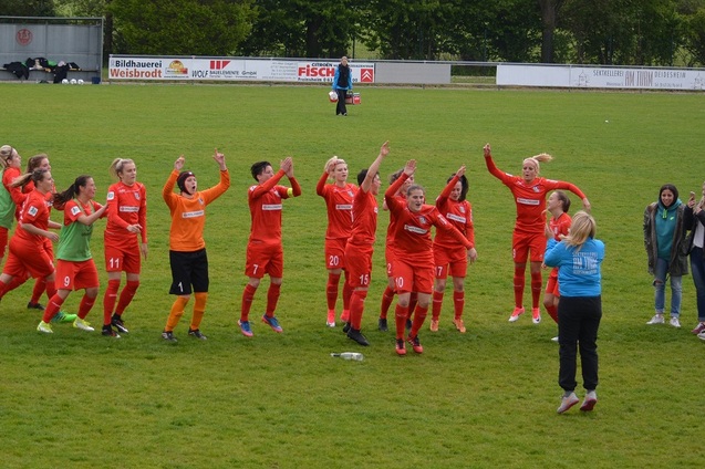 Frauenfu&szlig;ballmannschaft in roten Trikots jubelt auf dem Spielfeld, w&auml;hrend Zuschauer am Rand stehen.