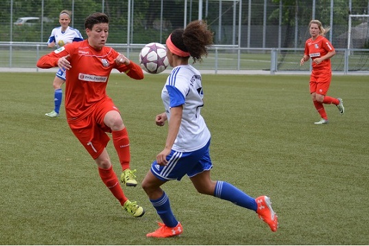 Spielszene eines Fu&szlig;ballspiels: Torh&uuml;terin mit rotem Trikot wehrt einen Schuss mit dem Kopf einer Spielerin im blauen Trikot ab.