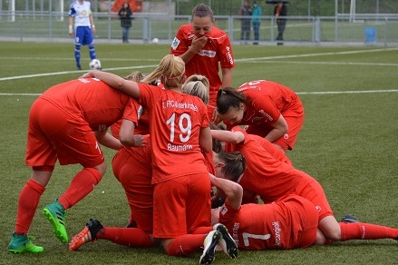 Gruppenbild von Spielerinnen in roten Trikots, die sich nach einem Torfeier auf einem Kunstrasenplatz umarmen.