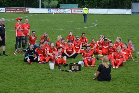 Gruppe von weiblichen Fu&szlig;ballspielerinnen in roten Trikots sitzt auf dem Rasen und freut sich gemeinsam.