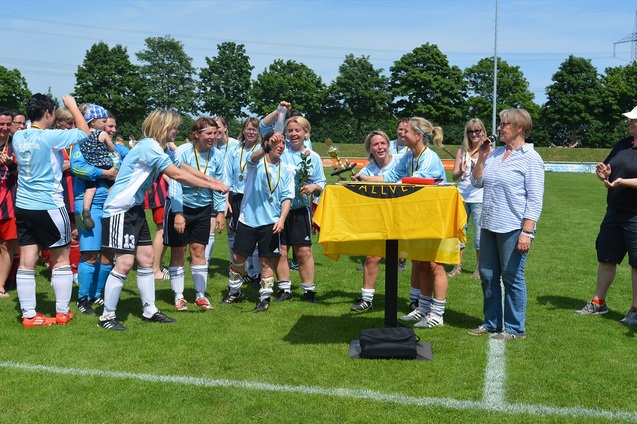 F&uuml;nfzehnfu&szlig;ballspielerinnen feiern um einen Tisch mit einer Fahne, w&auml;hrend ein Mitglied des Organisationsteams l&auml;chelt.