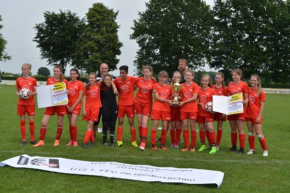 M&auml;dchenfu&szlig;ballmannschaft in roten Trikots h&auml;lt einen Pokal auf einem Spielfeld, umgeben von Betreuern und Plakaten.