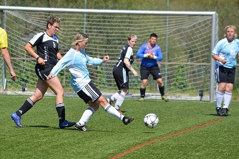 Zwei Frauen spielen Fu&szlig;ball auf einem Kunstrasenplatz, w&auml;hrend ein Torwart den Ball beobachtet.