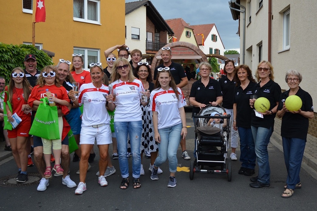 Gruppe von Menschen in sportlichen Outfits mit Sonnenbrillen, einige halten Getr&auml;nke und B&auml;lle, vor farbigen H&auml;usern.