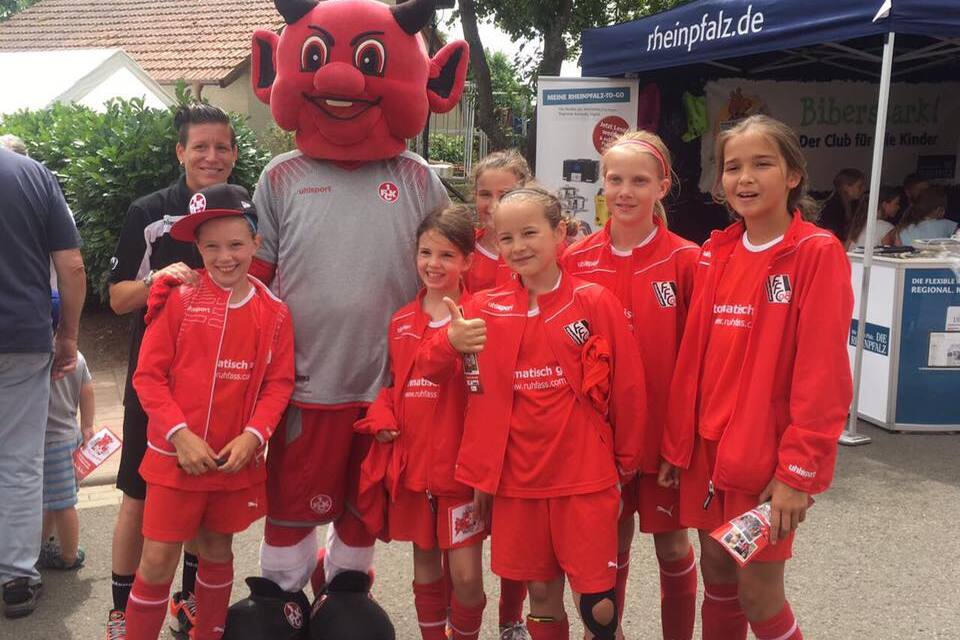 Gruppe von M&auml;dchen in roten Fu&szlig;balltrikots posiert mit einem Maskottchen auf einer Freiluftveranstaltung.