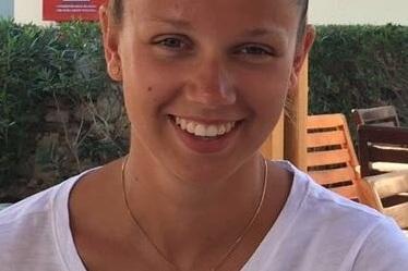 Lächelnde Person mit einem Dutt und Sonnenbrille sitzt an einem Tisch, im Hintergrund grüne Pflanzen und Holzstühle.