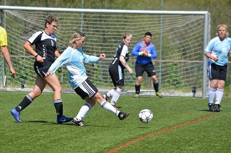 Zwei Frauen spielen Fu&szlig;ball auf einem Kunstrasenplatz, w&auml;hrend ein Torwart den Ball beobachtet.