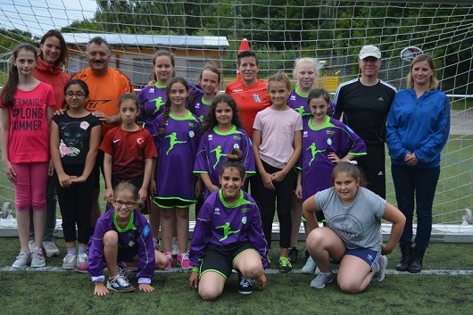Gruppenfoto von M&auml;dchen und Trainern in Sportkleidung vor einem Fu&szlig;balltor auf einem Rasenplatz.