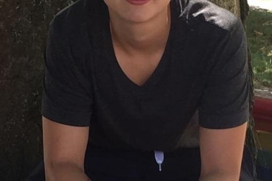 Junge Person mit kurzem Haar sitzt entspannt auf einer Bank vor einem Baum und l&auml;chelt in die Kamera.