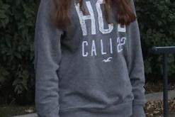 Junge Frau mit langen Haaren steht lächelnd im Freien, trägt einen grauen Pullover und Jeans. Hintergrund mit Bäumen.