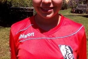 Mädchen im roten Fußballtrikot des FC Freiburg steht lächelnd auf einer Wiese, im Hintergrund Bäume und Möbel.