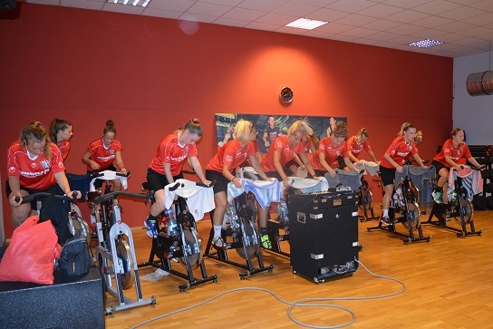 Gruppentraining auf Spinning-Bikes mit Frauen in roten Trikots in einem Fitnessraum mit Holzboden.