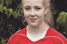 Portr&auml;t eines jungen M&auml;dchens mit blonden Haaren in rotem Fu&szlig;balltrikot vor gr&uuml;nen Pflanzen.