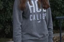 Junge Frau mit langen Haaren steht l&auml;chelnd im Freien, tr&auml;gt einen grauen Pullover und Jeans. Hintergrund mit B&auml;umen.