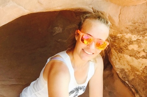 Frau mit Sonnenbrille l&auml;chelt in einer H&ouml;hle aus rotem Felsen und tr&auml;gt ein &auml;rmelloses Shirt und Shorts.