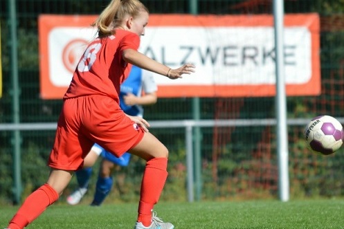 M&auml;dchen in rotem Fu&szlig;balltrikot schie&szlig;t einen Ball auf einem gr&uuml;nen Rasenplatz, im Hintergrund Banner sichtbar.