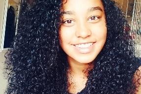 Lächelnde Person mit lockigem, schwarzem Haar vor einem hellen Hintergrund.