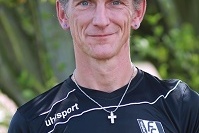 Portr&auml;t eines Mannes in schwarzem Sportshirt mit Kreuzkette, Hintergrund mit Pflanzen.