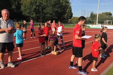 Gruppe von Kindern in roten T-Shirts steht auf einer Laufbahn; ein Erwachsener beobachtet die Aktivit&auml;ten.