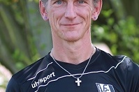 Portr&auml;t eines Mannes in schwarzem Sportshirt mit Kreuzkette, Hintergrund mit Pflanzen.