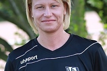 Portr&auml;t einer blondierten Frau in schwarzem Fu&szlig;balltrikot vor einer Pflanzenkulisse. Sie l&auml;chelt freundlich in die Kamera.