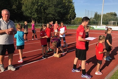Gruppe von Kindern in roten T-Shirts steht auf einer Laufbahn; ein Erwachsener beobachtet die Aktivit&auml;ten.