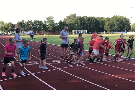 Gruppe von Kindern startet einen Lauf auf einer Sportbahn, betreut von einem Erwachsenen im Hintergrund.