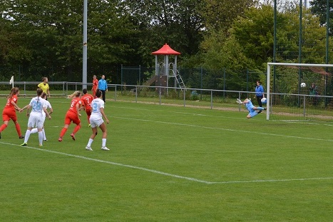Spielszene: Eine Spielerin tritt einen Elfmeter, w&auml;hrend die Torh&uuml;terin zur Abwehr springt. Im Hintergrund ist ein Sportplatz sichtbar.