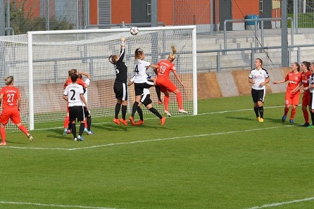 Spielszene im Frauenfu&szlig;ball: Torwart springt, um einen Ball w&auml;hrend eines Freisto&szlig;es abzuwehren, umgeben von Spielerinnen.