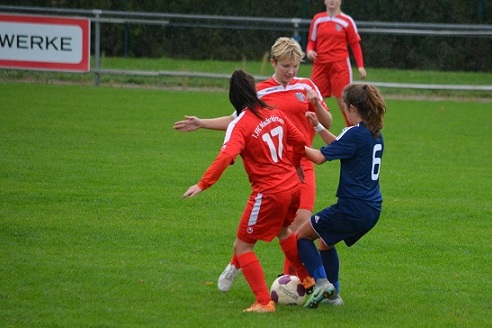 Zwei Fu&szlig;ballspielerinnen im Wettkampf um den Ball auf einem gr&uuml;nen Spielfeld, eine in Rot, eine in Blau.