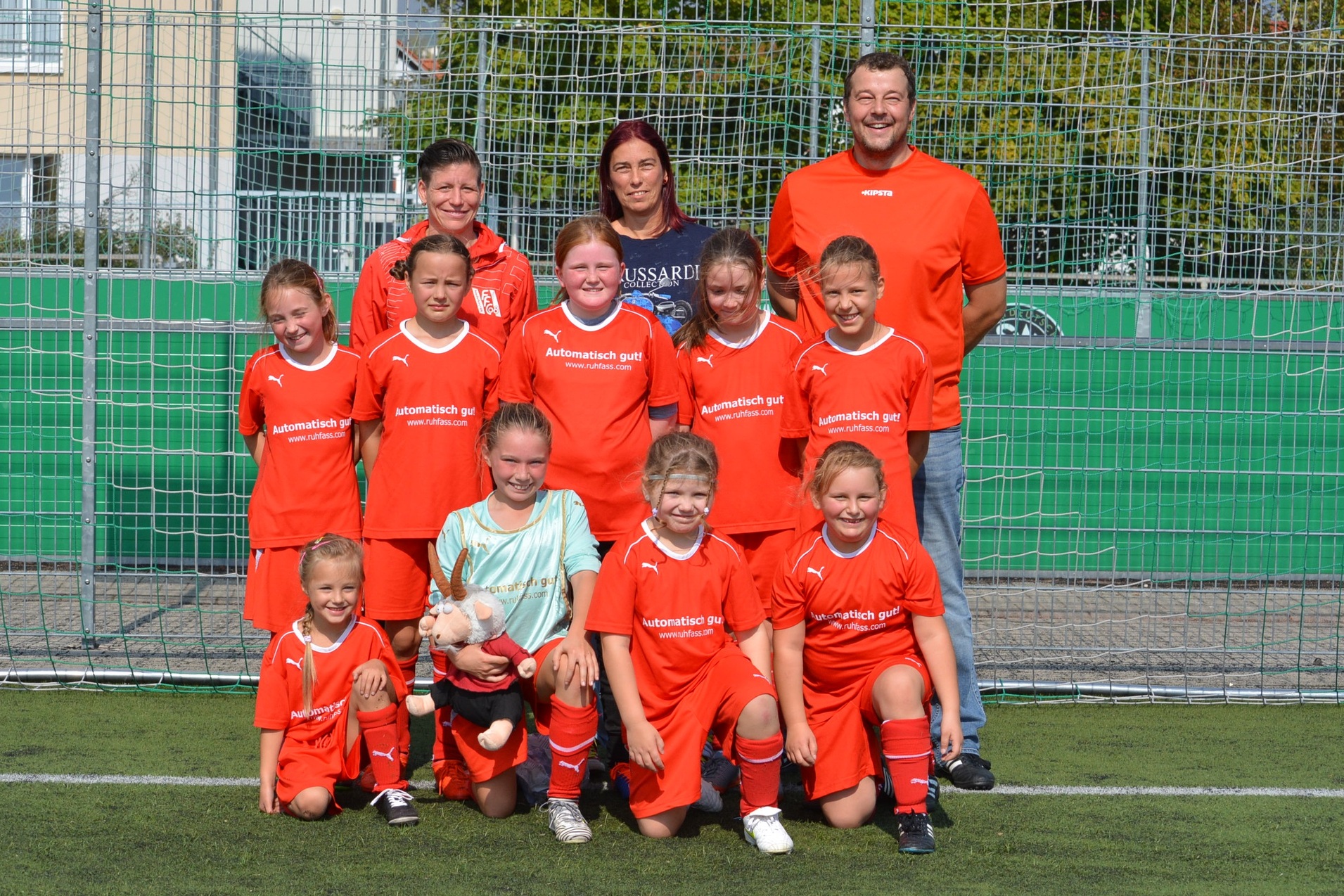 Gruppenfoto einer Jugendfu&szlig;ballmannschaft in orangen Trikots, begleitet von zwei Trainern auf einem Kunstrasenplatz.