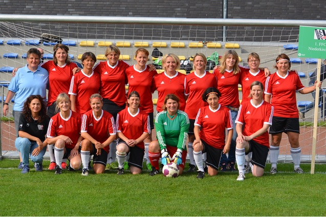 Gruppensfoto einer Frauenfu&szlig;ballmannschaft in roten Trikots vor einem Tor, mit einer Torfrau in gr&uuml;n und einem Vereinsbanner.
