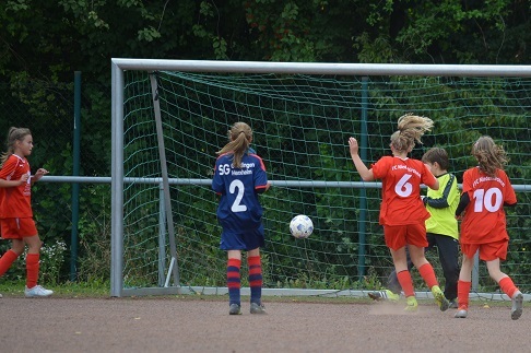 M&auml;dchen in roten Trikots und ein M&auml;dchen in Blau spielen Fu&szlig;ball, ein Ball fliegt ins Tor.