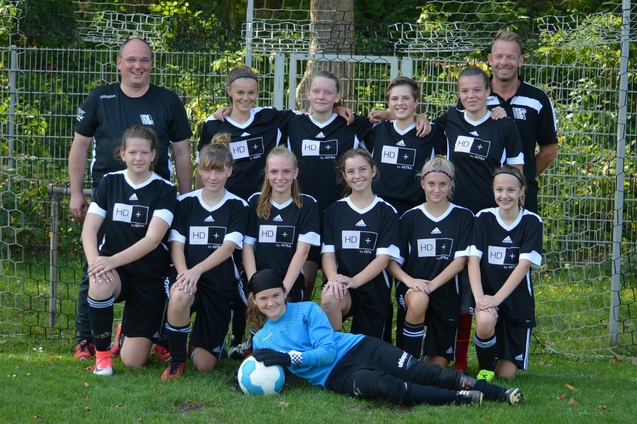 M&auml;dchenfu&szlig;ballmannschaft posiert in einheitlichen Trikots vor einem Fu&szlig;balltor mit zwei Trainern im Hintergrund.