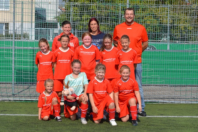 Gruppenfoto einer Jugendfu&szlig;ballmannschaft in orangen Trikots, begleitet von zwei Trainern auf einem Kunstrasenplatz.