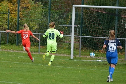 Torwartin in gr&uuml;ner Ausr&uuml;stung springt, w&auml;hrend eine Spielerin in rot auf das Tor zul&auml;uft. Fu&szlig;ball rollt ins Netz.
