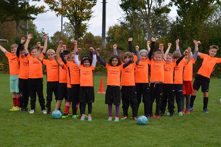 Gruppe von Kindern in orangefarbenen T-Shirts, die auf einem Fußballfeld jubeln, mit Fußbällen und Markierungsgegenständen.