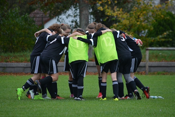 M&auml;dchenfu&szlig;ballmannschaft in Schwarz und Gr&uuml;n bildet einen Kreise auf dem Gras, H&auml;nde aufeinander gelegt f&uuml;r ein Teamritual.