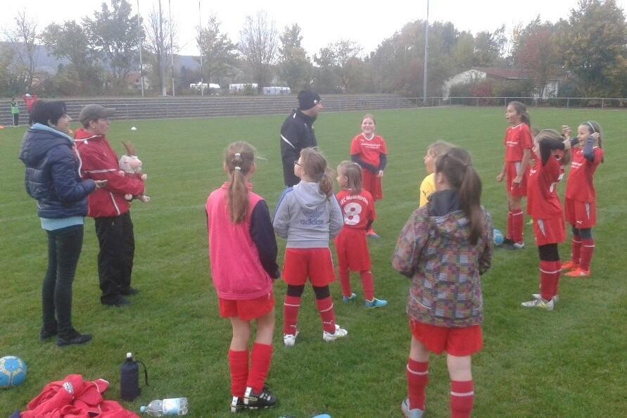 Gruppentraining eines Mädchenteams auf einem Fußballplatz, Trainer spricht mit den Spielerinnen in roten Trikots.