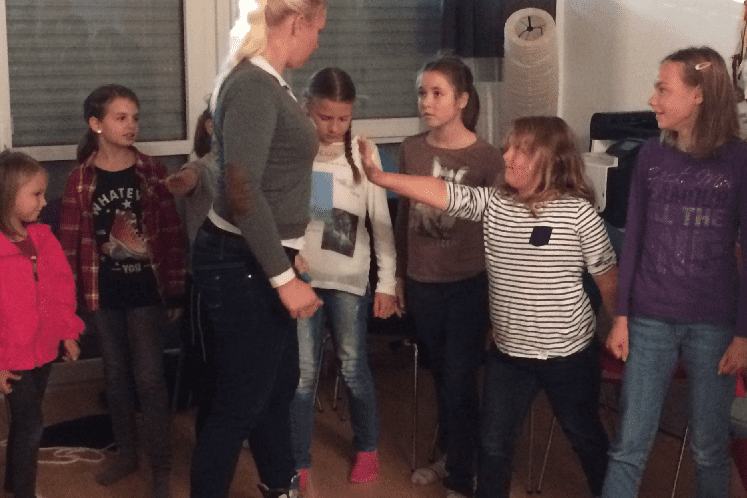 Gruppenszene mit acht M&auml;dchen, die einer stehenden Trainerin in einem Raum aufmerksam zuh&ouml;ren.