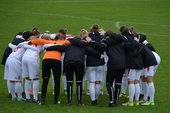 Frauenfu&szlig;ballmannschaft in geschlossener Formation, die sich umarmt und bespricht, auf einem gr&uuml;nen Spielfeld.