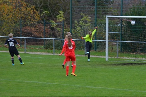 Spielerinnen in roten und schwarzen Trikots; eine Torh&uuml;terin springt, um einen Ball zu halten, der in Richtung Tor fliegt.