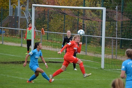 Torh&uuml;terin in orange springt, um einen Ball auf dem Fu&szlig;ballplatz zu parieren, w&auml;hrend Spielerinnen in blauen und gr&uuml;nen Trikots agieren.