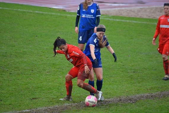 Zwei Fu&szlig;ballspielerinnen im Wettkampf um den Ball auf einem matschigen Spielfeld. Eine Spielerin in Rot, die andere in Blau.