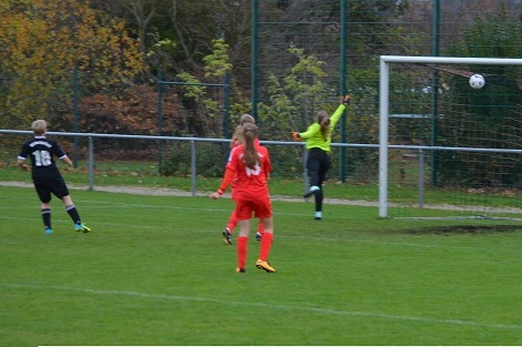 Spielerinnen in roten und schwarzen Trikots; eine Torh&uuml;terin springt, um einen Ball zu halten, der in Richtung Tor fliegt.