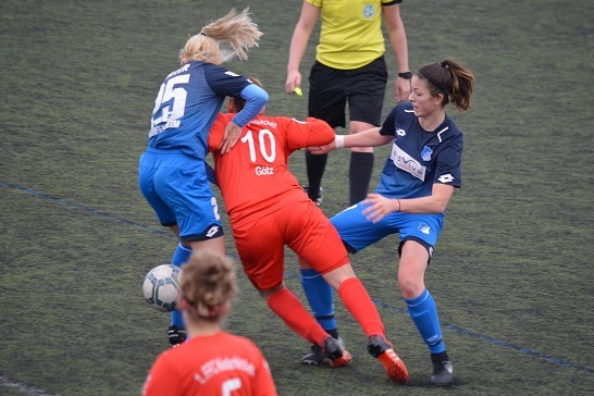 Zwei Spielerinnen im Zweikampf, eine in roter und eine in blauer Kleidung, auf einem Fu&szlig;ballfeld. Schiedsrichterin im Hintergrund.