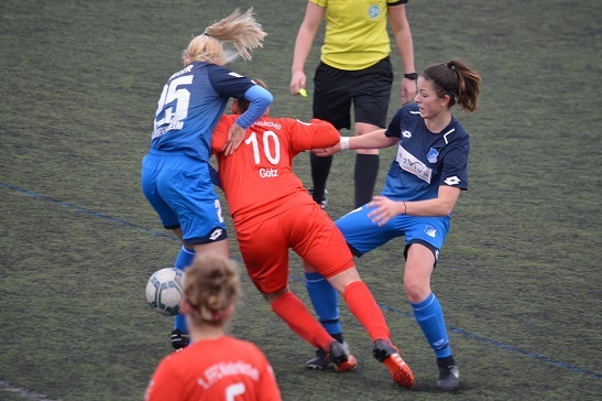 Zwei Spielerinnen im Zweikampf, eine in roter und eine in blauer Kleidung, auf einem Fu&szlig;ballfeld. Schiedsrichterin im Hintergrund.