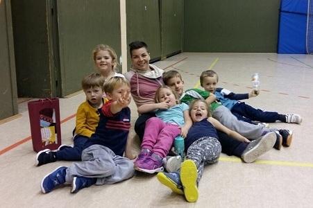 Gruppe von acht Kindern und einem Erwachsenen, die auf dem Boden einer Sporthalle sitzen und fr&ouml;hlich posieren.
