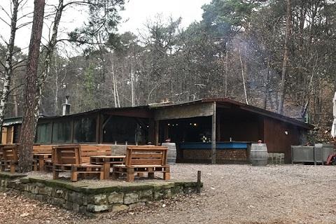 Holzh&uuml;tte im Wald mit Au&szlig;enterrasse, Tischen und B&auml;nken, umgeben von B&auml;umen und leichtem Rauch.